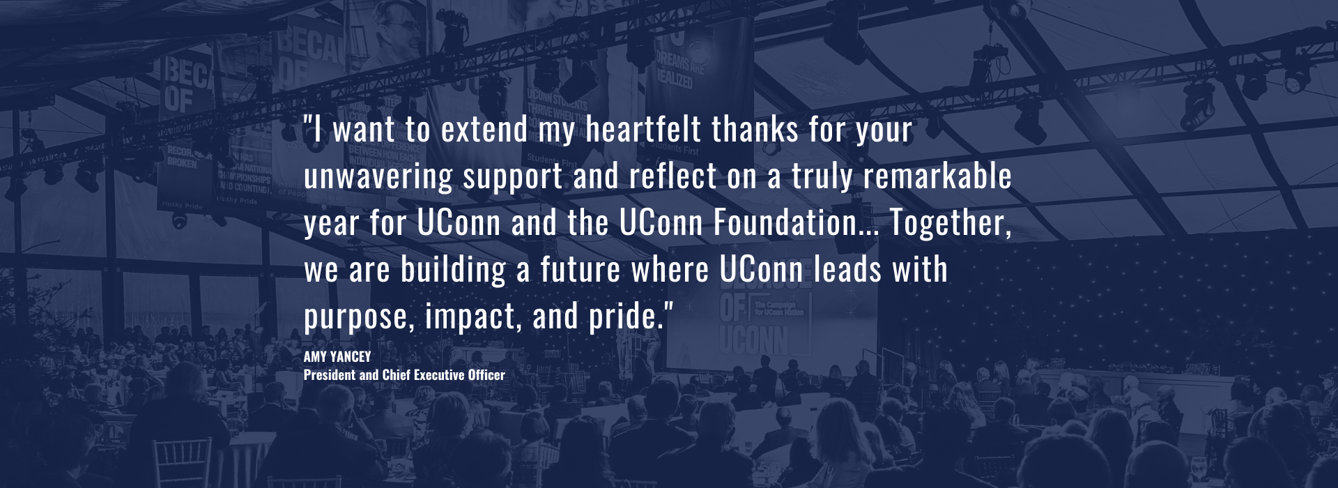 "I want to extend my heartfelt thanks for your unwavering support and reflect on a truly remarkable year for UConn and the UConn Foundation... Together, we are building a future where UConn leads with purpose, impact, and pride." Amy Yancey, President and Chief Executive Officer, background picture is a room full of seated people looking at a man and a woman standing on a stage at a podium in front of a screen that says "Because of UConn"